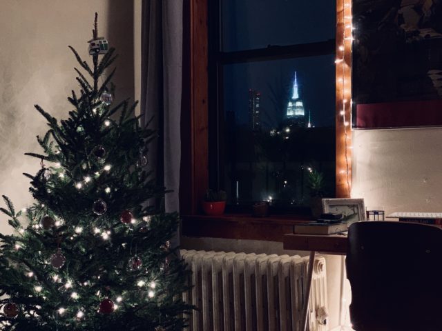 Empire State é visto da janela do apartamento em Manhattan. O edifício tem uma antena pontuda em seu topo e está iluminado de branco. Ao lado da janela há uma árvore de natal.
