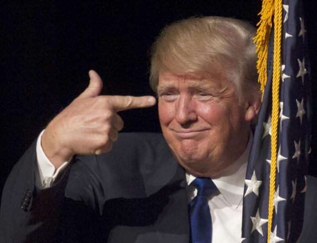 Donald Trump apontando para a própria cabeça com a mão em forma de revólver, como quem simboliza um suicídio