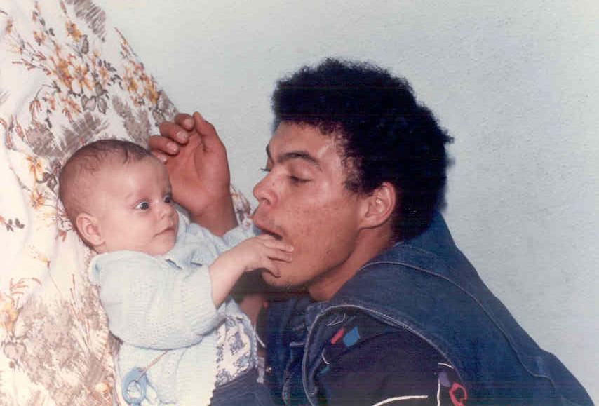 Marco Gomes, bebê, com os olhos arregalados, coloca a mão dentro da boca do pai, que brinca de morder seus dedos. Wilian Gomes tem cabelo black power curto, e várias marcas de espinhas no rosto.