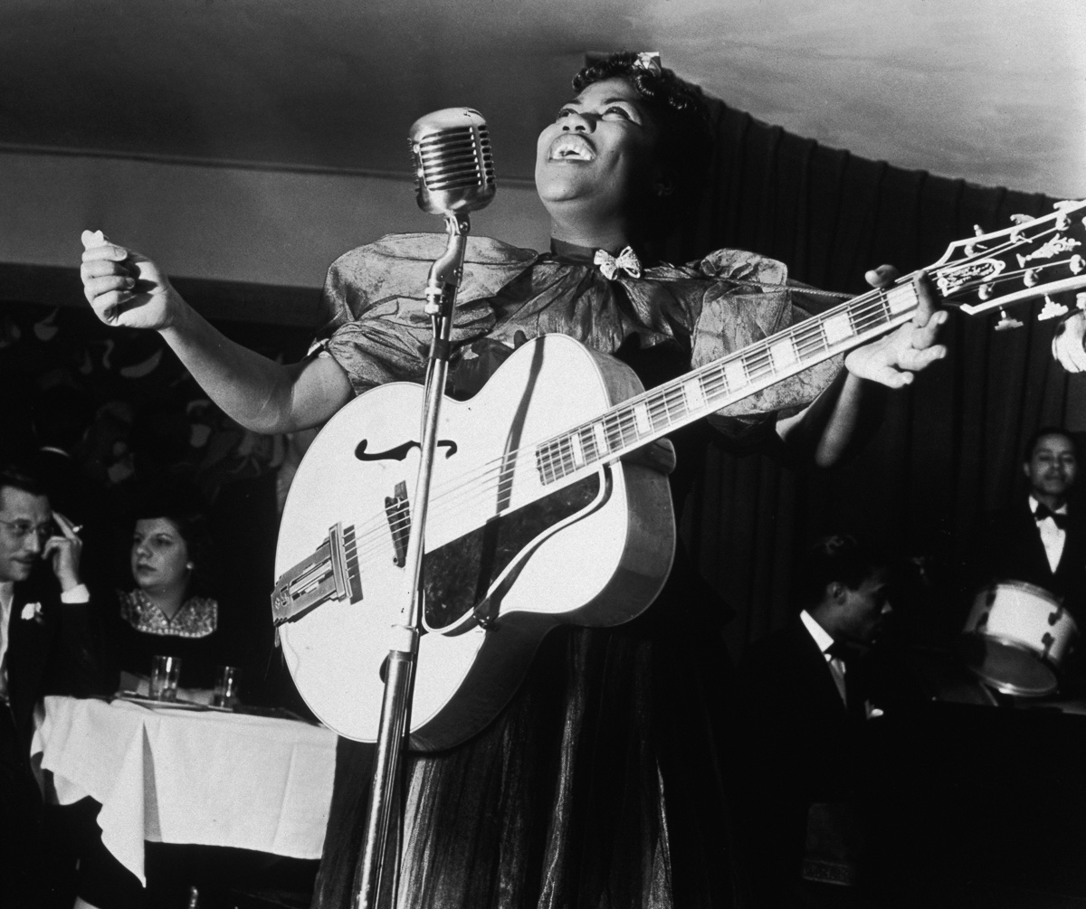 Sister Rosetta Tharpe, 1938. Uma mulher preta e gorda tocando guitarra e cantando em um salão de festas, atrás dela à esquerda da imagem, um casal sentado à mesa, atrás dela à direita da imagem, músicos da banda que a acompanhava.