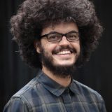 Marco Gomes em 2022, sorri largamente, cabelo afro, óculos, barba cheia, camisa em padrão xadrez cinza escuro e verde, abotoada até o pescoço.