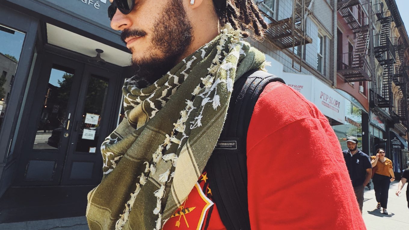 Marco Gomes caminha em uma rua urbana ensolarada. Ele usa óculos escuros, fones de ouvido e veste uma camisa vermelha com detalhes pretos e amarelos, além de um cachecol tipo keffiyeh verde. Seu cabelo está em dreadlocks, preso por uma faixa preta. Ao fundo, há prédios típicos com escadas de incêndio e uma fachada de café com a placa “Hungry Coffee Bar & Café”.