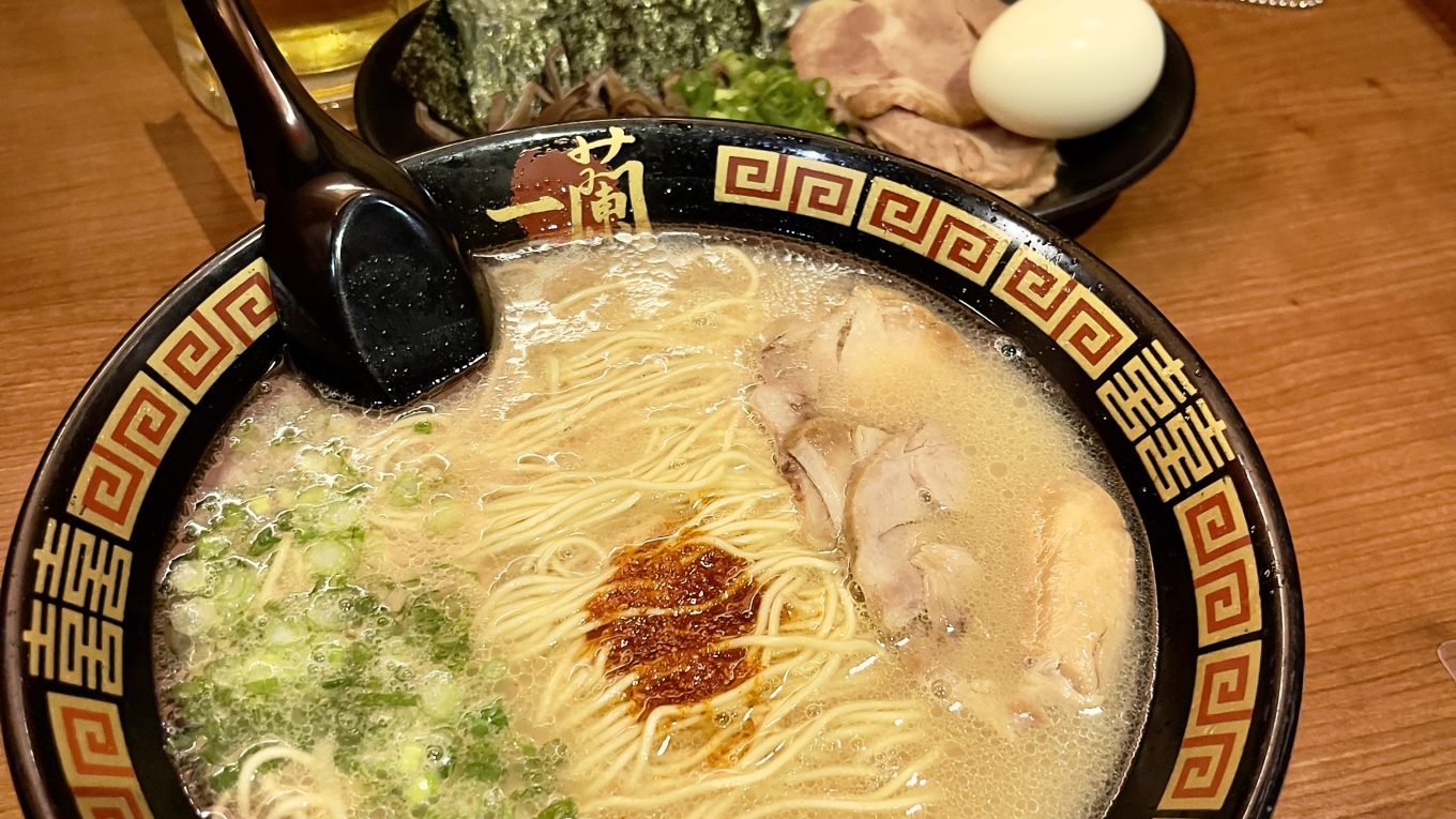 A imagem mostra uma tigela de tonkotsu ramen do Ichiran, com caldo cremoso, noodles, carne de porco, cebolinha e uma porção de molho picante no centro. Ao lado, há um prato com um ovo cozido, algas nori e fatias adicionais de carne de porco. Atrás, um copo de cerveja “Super Dry” completa a refeição, servida em uma cabine de madeira aconchegante.