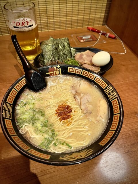 A imagem mostra uma tigela de tonkotsu ramen do Ichiran, com caldo cremoso, noodles, carne de porco, cebolinha e uma porção de molho picante no centro. Ao lado, há um prato com um ovo cozido, algas nori e fatias adicionais de carne de porco. Atrás, um copo de cerveja “Super Dry” completa a refeição, servida em uma cabine de madeira aconchegante.