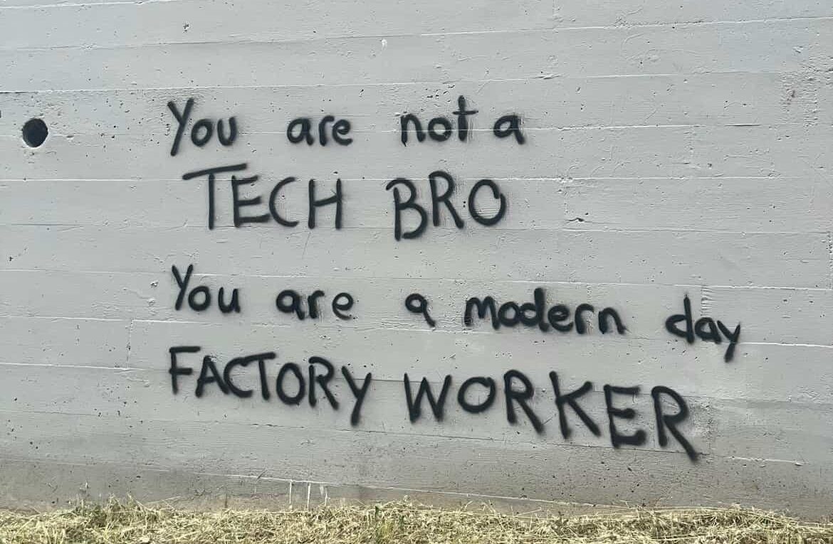 a imagem mostra um muro de concreto cinza, um pouco gasto, com grama seca na base. no centro, escrito em spray preto, aparece a frase “you are not a tech bro. you are a modern day factory worker”. em português: “você não é um tech bro. você é um trabalhador de fábrica dos dias atuais”. as letras são irregulares, feitas à mão, com estética de graffiti direto e crítico, como um recado para quem passa.