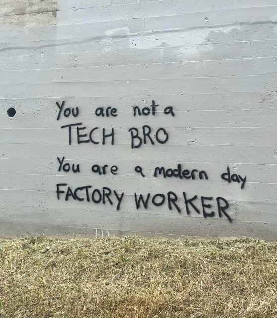 a imagem mostra um muro de concreto cinza, um pouco gasto, com grama seca na base. no centro, escrito em spray preto, aparece a frase “you are not a tech bro. you are a modern day factory worker”. em português: “você não é um tech bro. você é um trabalhador de fábrica dos dias atuais”. as letras são irregulares, feitas à mão, com estética de graffiti direto e crítico, como um recado para quem passa.
